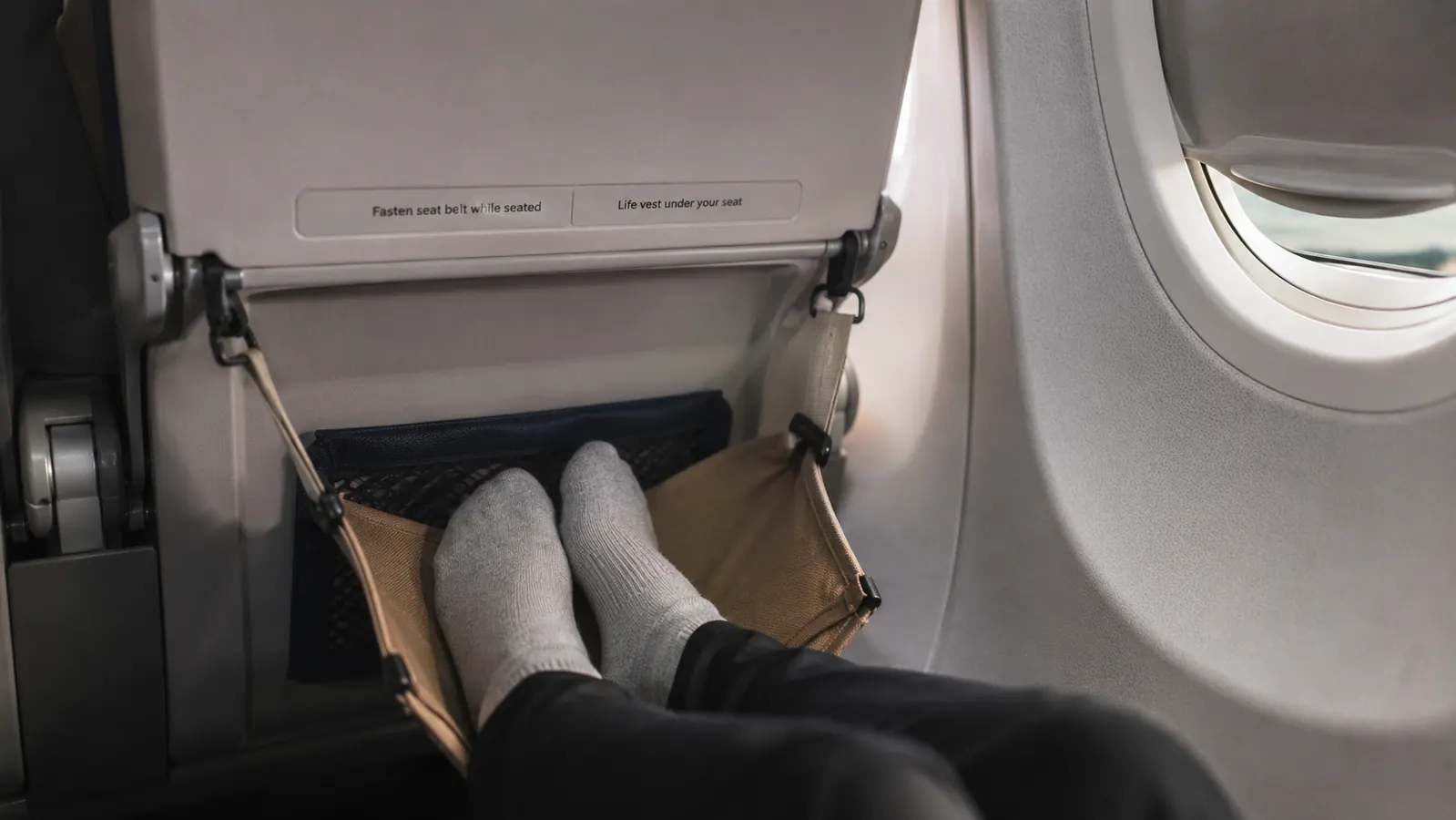 Traveler's feet resting elevated in a memory foam footrest hammock under an economy airplane seat
