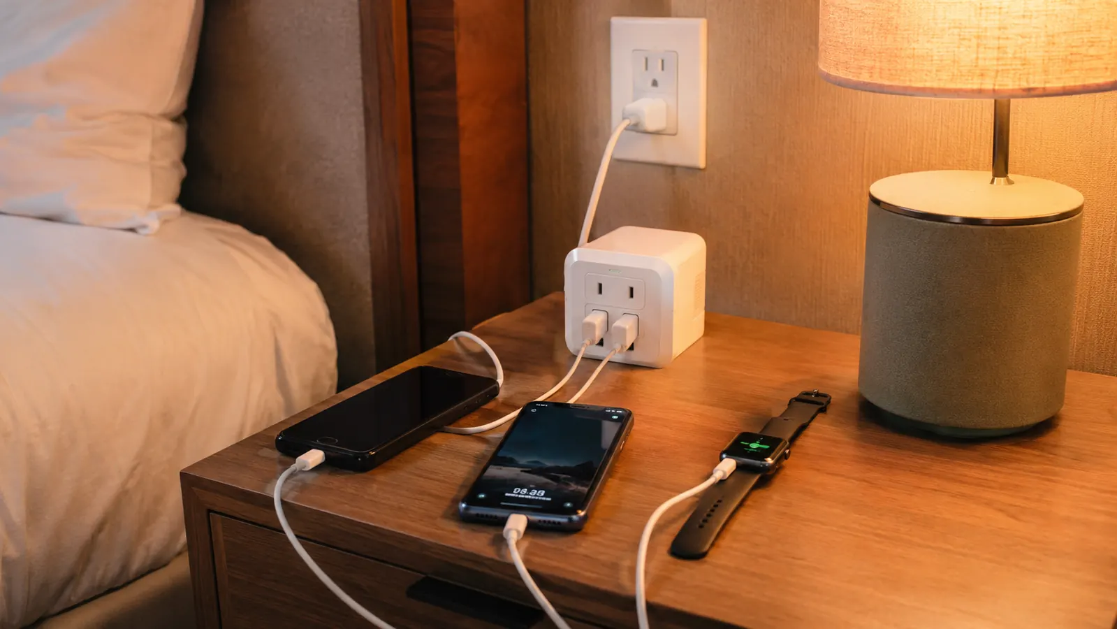 A compact power strip with multiple outlets and USB ports sitting on a hotel nightstand next to a lamp and two charging phones