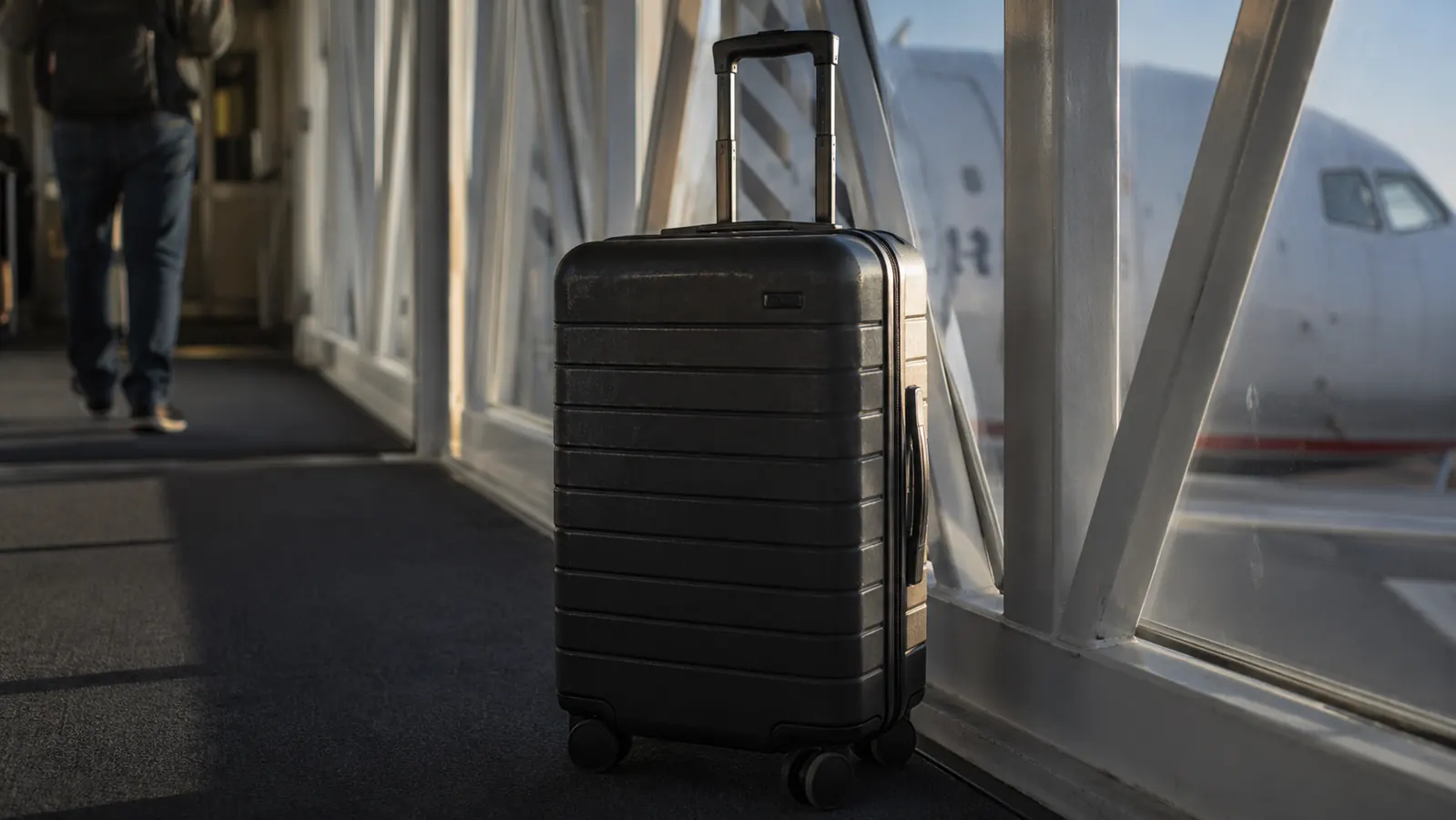 Black hardside carry-on suitcase sitting on a jet bridge floor with a scuffed corner visible in the light