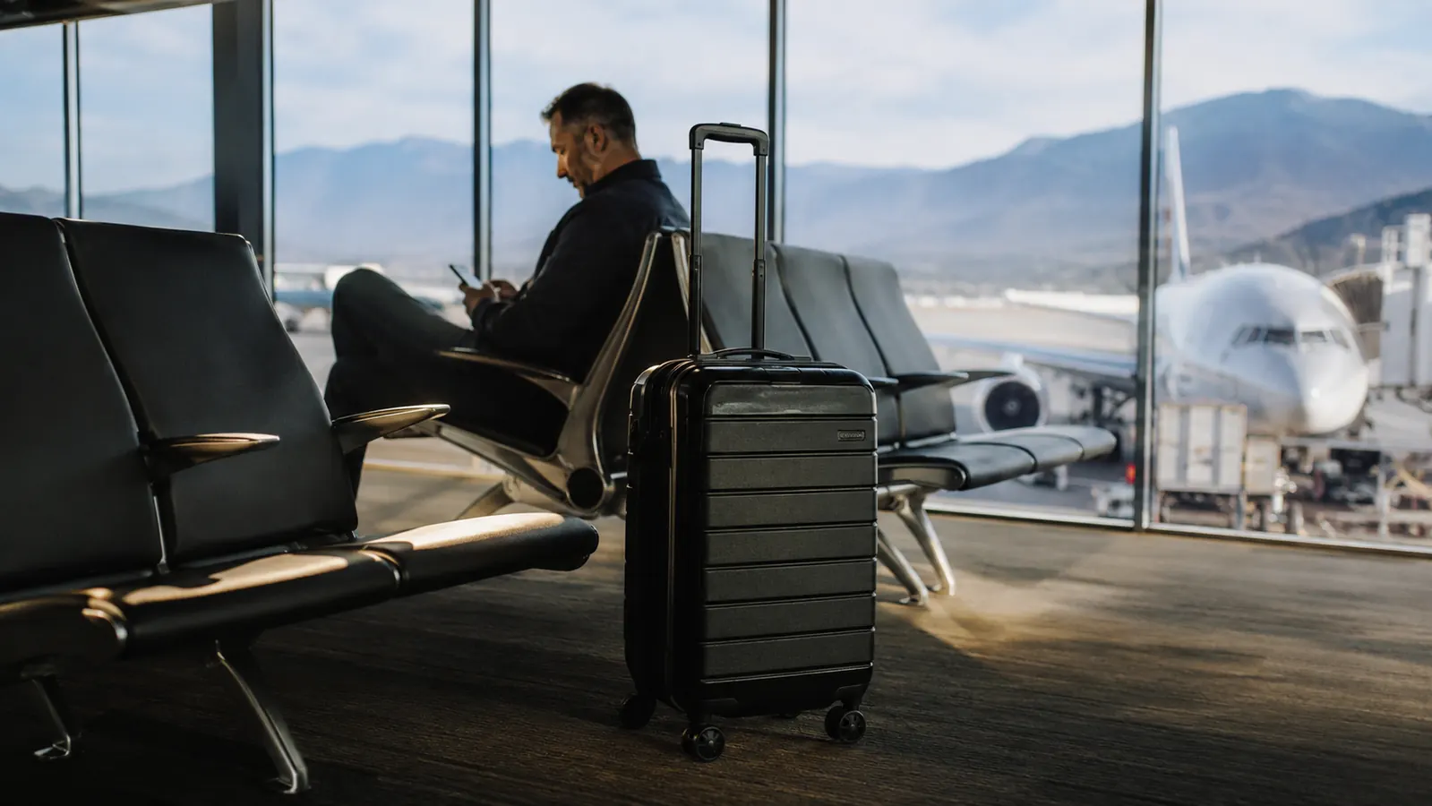 Black hardside carry-on suitcase standing upright at an airport gate near a row of windows