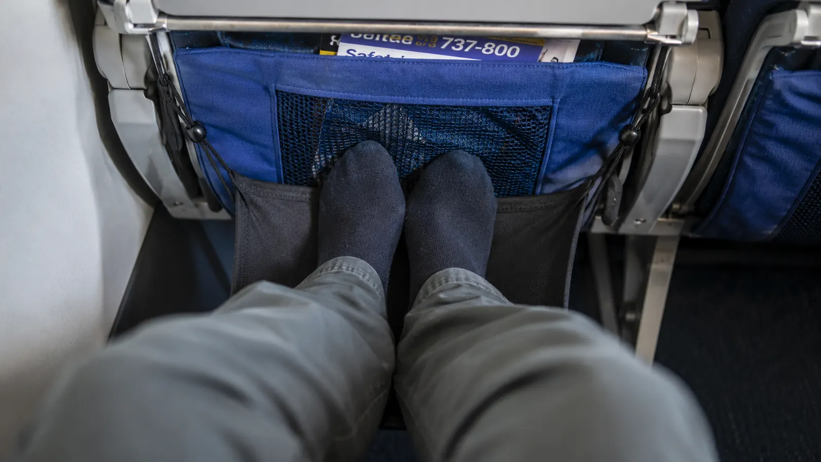 Airplane footrest hammock sagging slightly under a passenger's feet in a cramped economy seat