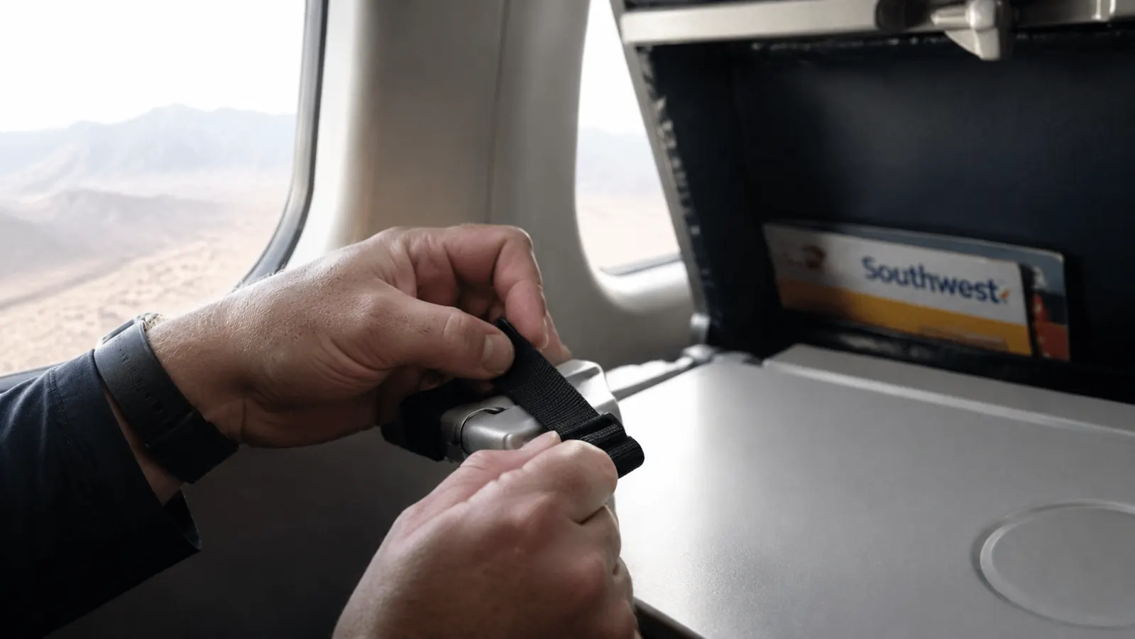 A traveler's hand adjusting the strap of a foot hammock looped around an airplane tray table arm