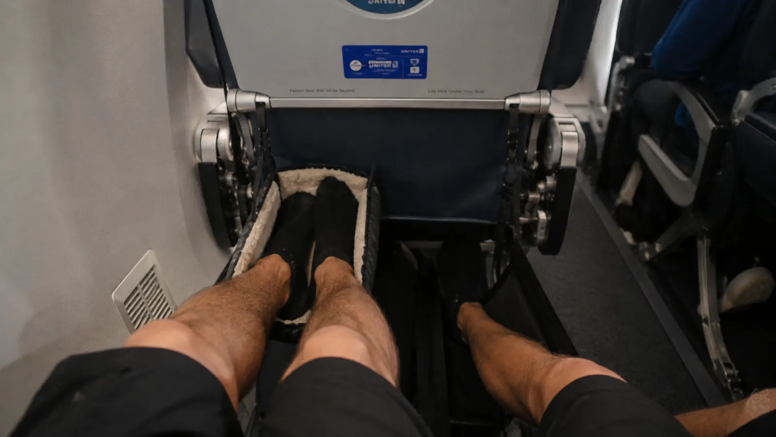 A padded memory foam airplane footrest and a plain canvas foot hammock hanging side by side from an airplane tray table