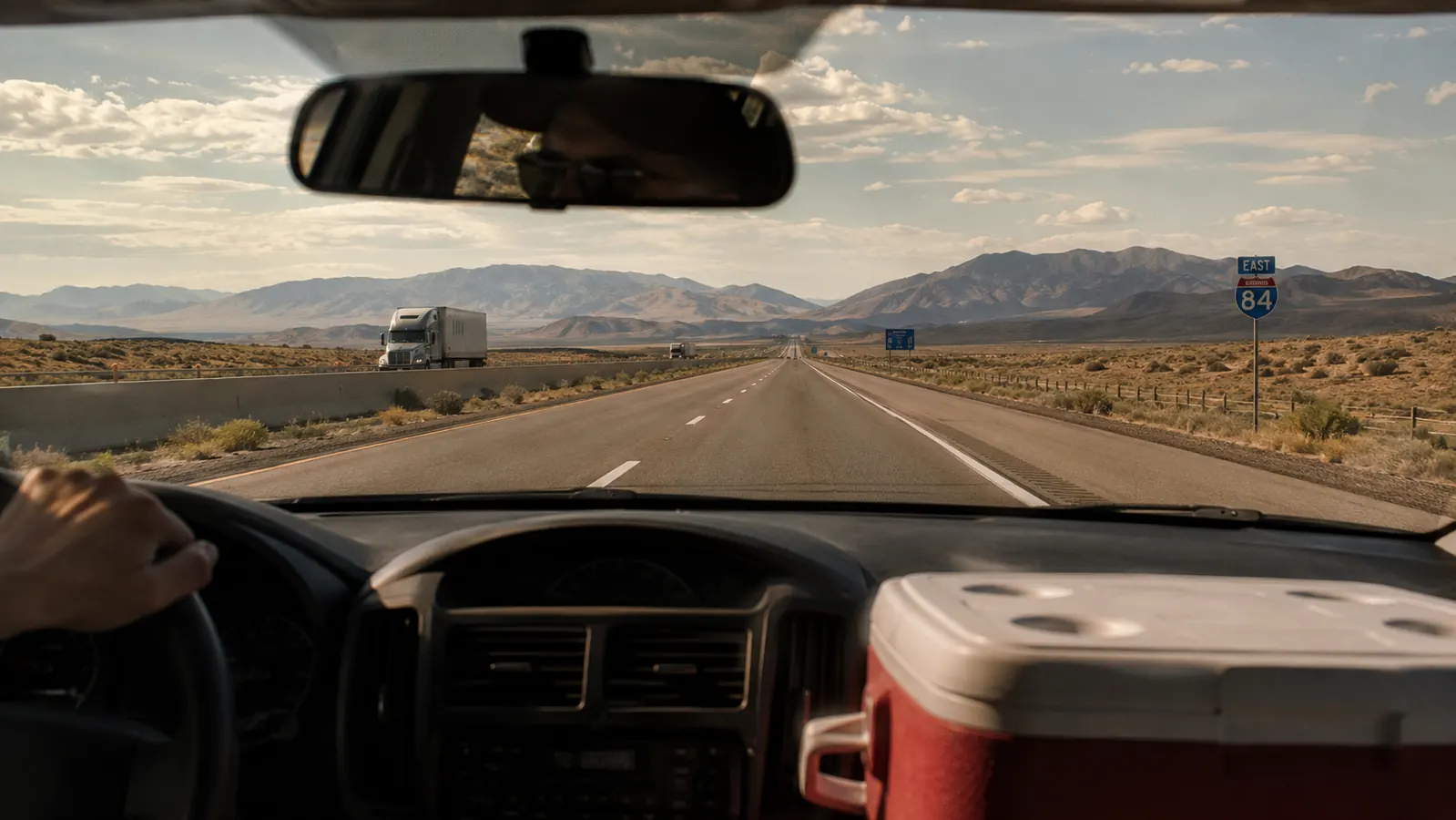 A dusty highway stretching through desert scrubland with a truck's dashboard and cooler visible in the foreground, heat shimmer on the road ahead