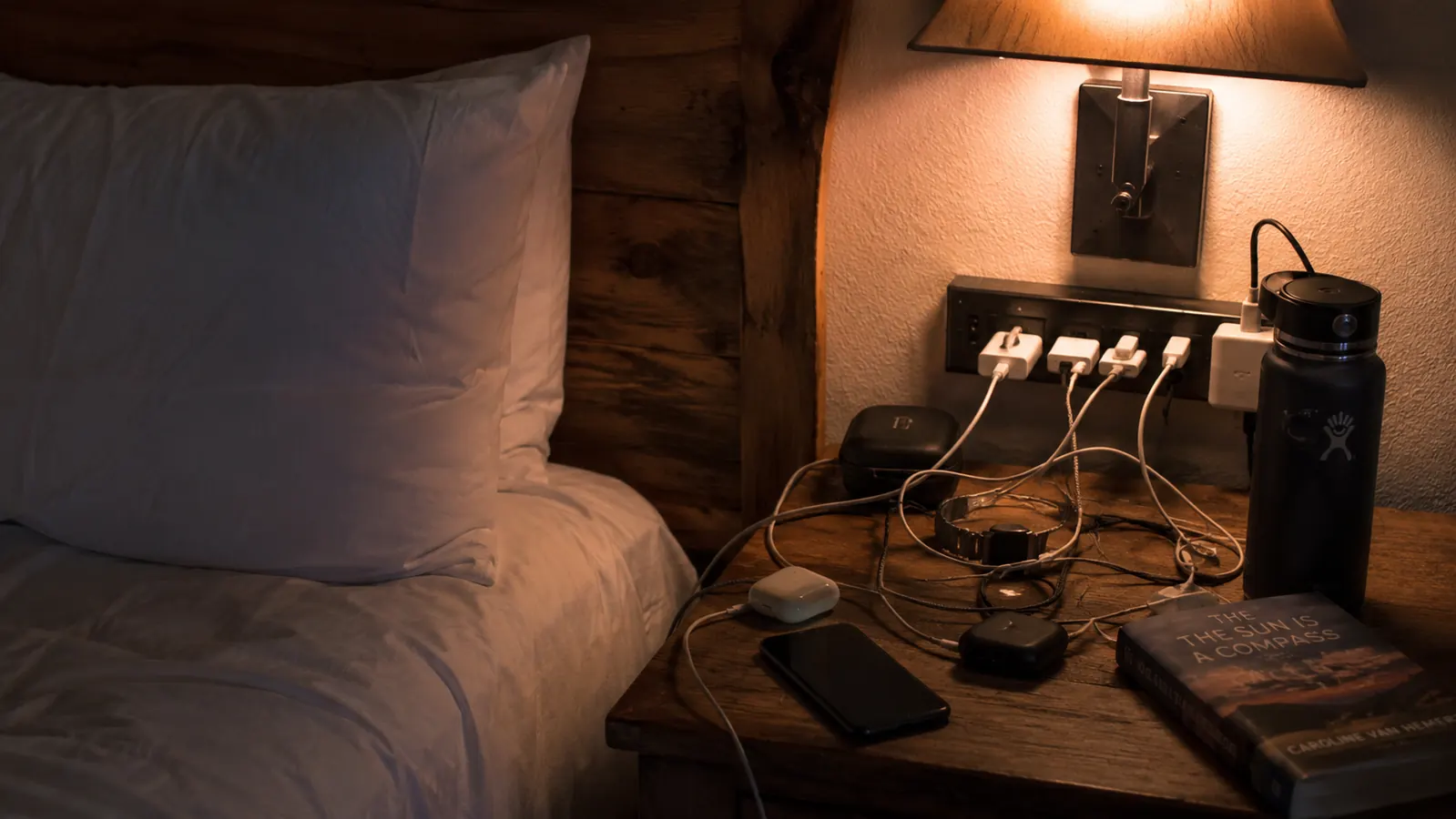 A hotel nightstand at night with a lamp, a phone charging cord, and a laptop cord all competing for a single wall outlet