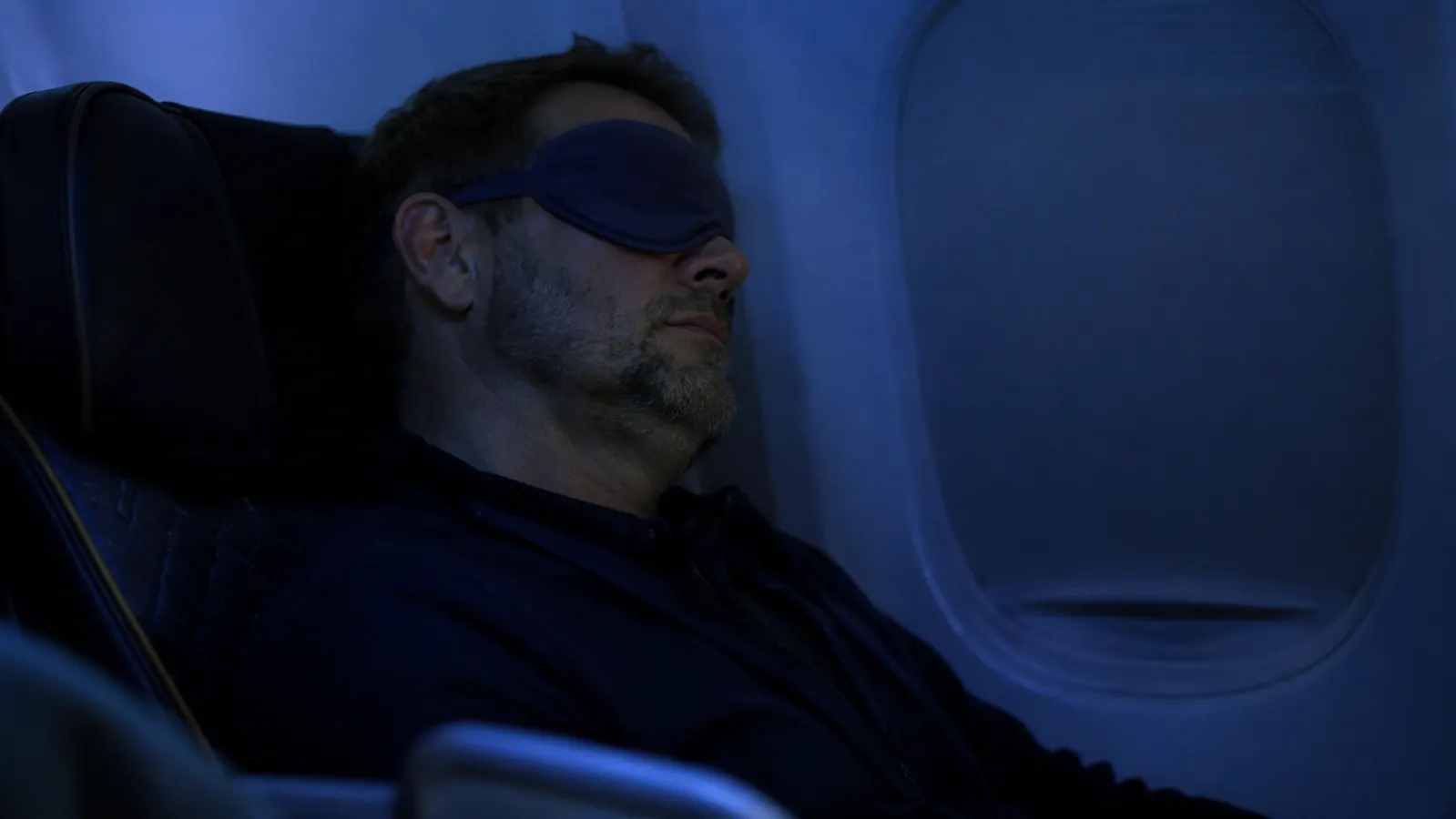 A traveler asleep in an economy airplane seat at night, cabin lights dimmed, window shade down