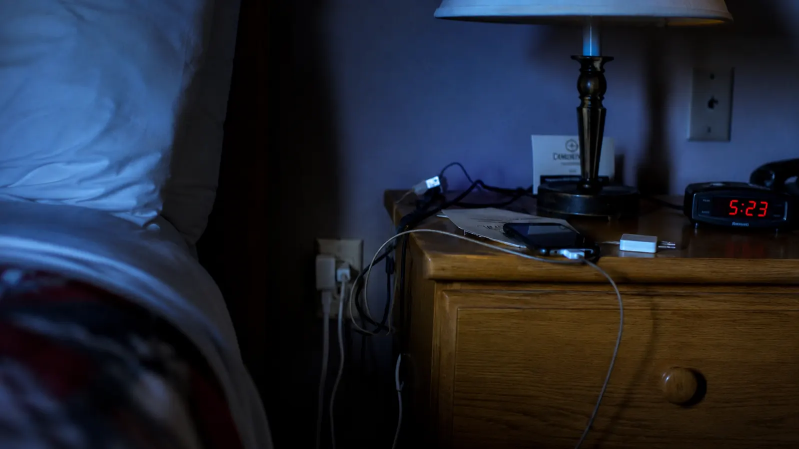 A dim motel nightstand with a lamp and a single crowded outlet behind it, early morning light through the curtains
