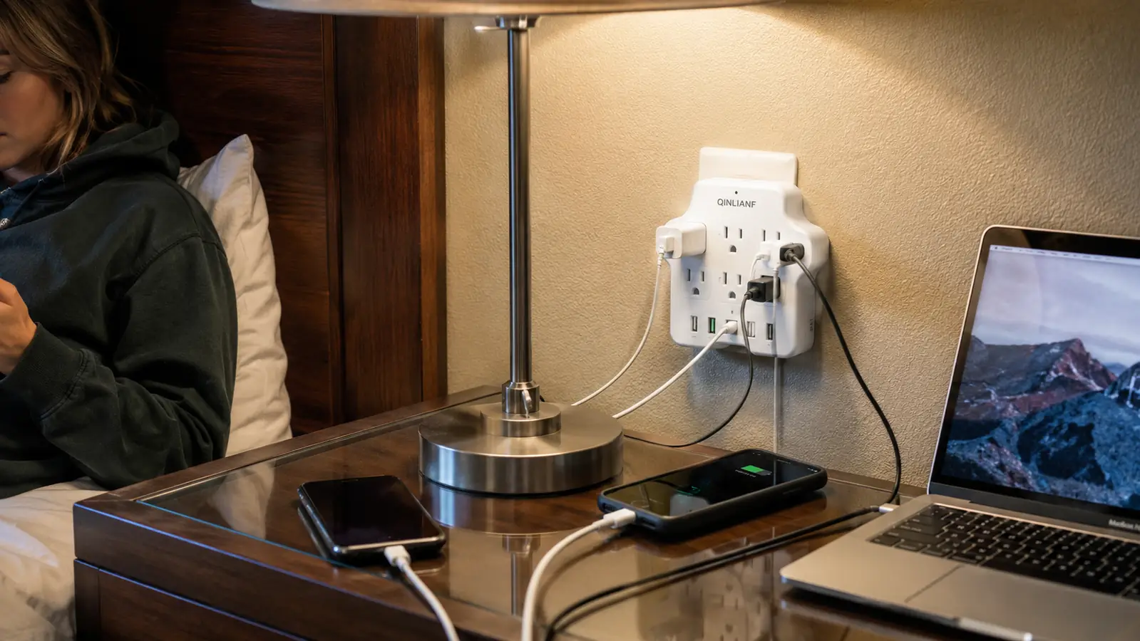 White travel power strip plugged into a hotel outlet behind a nightstand with two phones charging